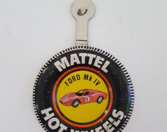 May include: A black and orange Hot Wheels button with a white background. The button features a red Ford MK IV race car on a yellow background. The text "MATTEL", "FORD MK IV", "HOT WHEELS", and "©1969 Mattel Inc. Hong Kong" is printed on the button.
