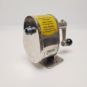 May include: A vintage Chicago No. 2 pencil sharpener with a metallic finish and a yellow label featuring addresses for the Woolworth Building, Grand Central, and Hotel Biltmore. It includes a side-mounted hand crank with a black handle knob.