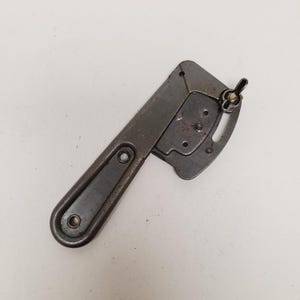 May include: A vintage metal tool featuring a long handle with two circular mounting holes and an adjustable head section. The head includes a curved slot and a metal wing nut fastener, showing signs of age with a dark, weathered gray finish.