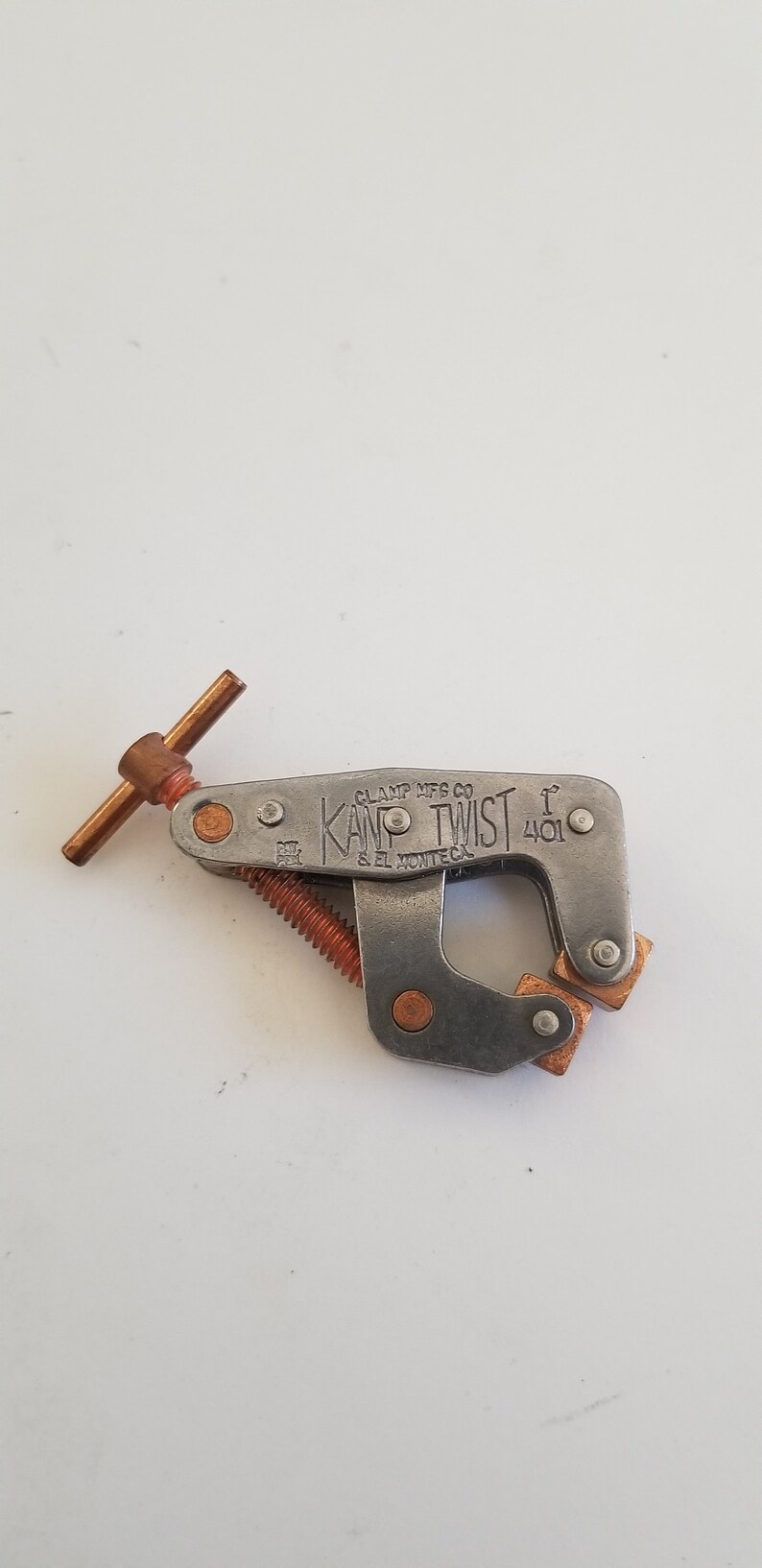 May include: A silver metal clamp with a copper-colored handle and a screw. The clamp is labeled "KAMP TWIST" and "CLAMP MFG CO. S. EL MONTE, CA. 401".
