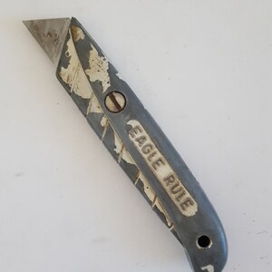 May include: A gray and white utility knife with a worn handle. The handle has the words "Eagle Rule" printed on it.