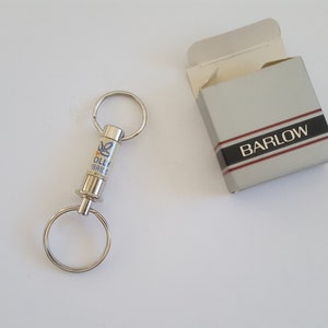 May include: A silver keychain with two rings and a cylindrical center, featuring blue and yellow text and a floral design. Beside it is a gray box with the word "BARLOW" in a black and red stripe.