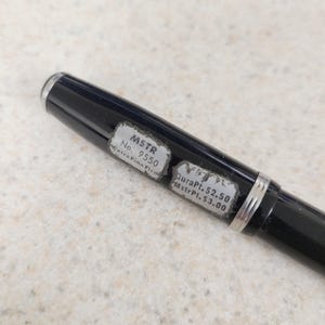 May include: A black pen with silver accents and a silver tip. The pen has two stickers with the text "MSTR No. 9550 Extra Fine Firm" and "DuraPt. $2.50 MstrPt. $3.00".
