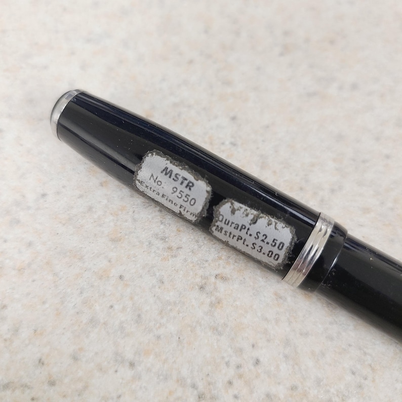 May include: A black pen with silver accents and a silver tip. The pen has two stickers with the text "MSTR No. 9550 Extra Fine Firm" and "DuraPt. $2.50 MstrPt. $3.00".