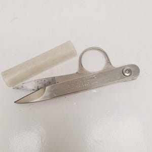 May include: A pair of Wiss-Clip scissors with a finger loop and a protective clear plastic sleeve. The scissors are made of silver-colored metal and have a pointed blade. The text "WISS-CLIP" is engraved on the scissors.
