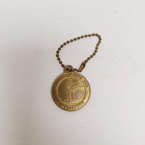 May include: A vintage brass-colored medallion with a ball chain. The medallion features an illustration of a person holding a staff, with the words "METHODIST MINUTE MEN STEWARDSHIP" around the edge. The chain is made of small, round beads.