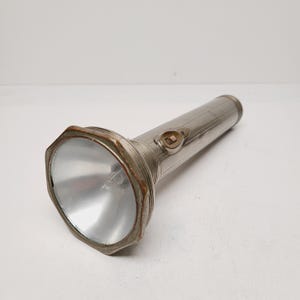 May include: A vintage silver flashlight with a large, octagonal head and a ribbed handle. The light has a clear lens and a small switch on the side. The metal shows signs of age and wear.