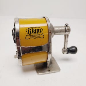 May include: Vintage yellow and silver pencil sharpener with the word "Giant" and the text "Made by Automatic Pencil Sharpener Co. Chicago, U.S.A." printed on the side. The sharpener has a crank handle with a black knob.