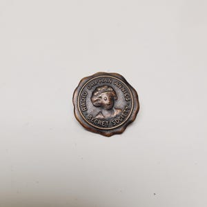 May include: A circular, bronze-colored pin featuring a relief of a girl's face and the text "Radio Orphan Annie's Secret Society" around the edge. The pin has a slightly raised border and a vintage aesthetic.