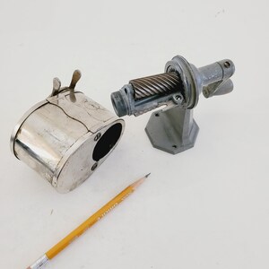 Puede incluir: Un sacapuntas vintage de plata con un contenedor de metal separado para virutas. El sacapuntas tiene una manivela y una cuchilla de metal. Un l&aacute;piz amarillo est&aacute; sobre la superficie blanca.