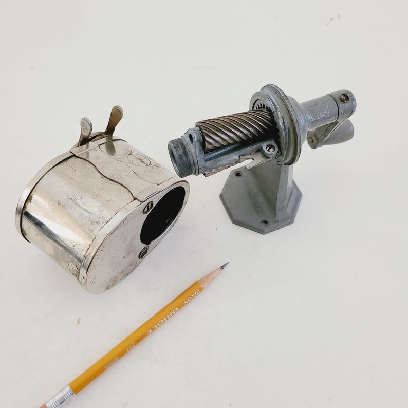 Puede incluir: Un sacapuntas vintage de plata con un contenedor de metal separado para virutas. El sacapuntas tiene una manivela y una cuchilla de metal. Un l&aacute;piz amarillo est&aacute; sobre la superficie blanca.