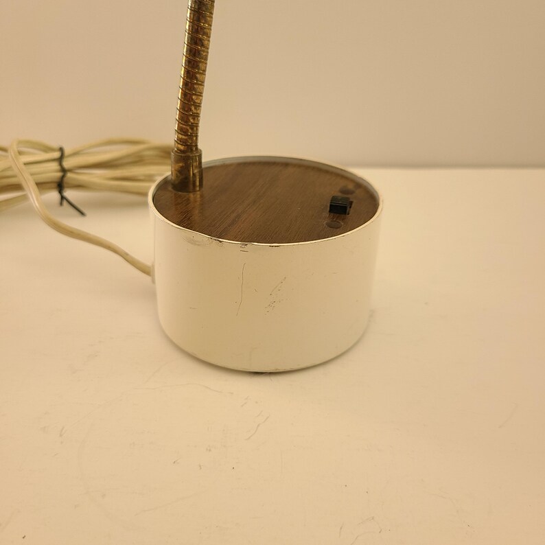 May include: A small, white desk lamp with a flexible, brass-colored neck. The lamp has a round, wooden top and a white cylindrical base. A power cord extends from the base.