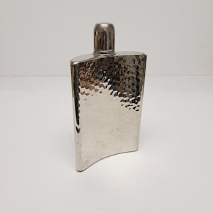 May include: A silver-colored metal flask with a curved, rectangular body and a hammered texture finish. The flask features a smooth, cylindrical screw-top cap at the center of the top edge, reflecting light across its metallic surface.