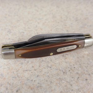 May include: A closed folding pocket knife with a brown, textured handle and silver-colored metal bolsters at both ends. The handle features an oval silver inlay engraved with the text L.L. Bean. The knife has two folded steel blades.