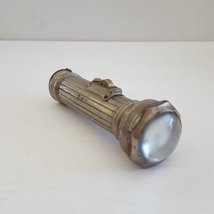 May include: A vintage silver flashlight with a clear, rounded lens. The flashlight has a ribbed body and a switch on top. The design is classic and functional, suggesting a focus on utility and durability.
