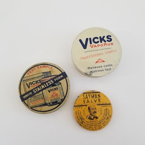 May include: Three vintage tin containers for ointment. The largest container is white with blue lettering and reads "Vicks VapoRub Vaporizing Ointment Professional Sample Relieves colds distress fast". The medium container is yellow with red and black lettering and reads "Vicks VapoRub White Stainless Form For Greasy Treatment of INFLAMMATION CONGESTION". The smallest container is yellow with black lettering and reads "Sayman Salve Net Wt. Not Over 1/4 Oz. Free Sample Trade Mark Ingredients Camphor, Oxide Zinc, Boric Acid, Lanolin Petroleum For Many Applications and Protective Dressing Irritations of the Skin and Scalp Sayman Products Co. St. Louis, Mo."