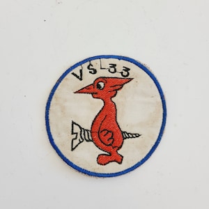 May include: A white, round patch with a blue border. The patch features a red cartoon bird with a screw sticking out of its back. The text "VS 33" is embroidered in black above the bird.