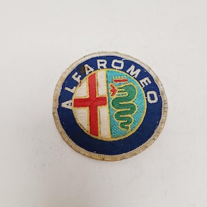 May include: Circular Alfa Romeo patch with a blue border. The patch features a red cross on a white background and a green serpent with a crown on a light blue background. The words "ALFAROMEO" are embroidered around the edge.