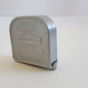 Puede incluir: Un flex&oacute;metro de metal plateado con cuerpo rectangular y parte superior redondeada. El texto "MADE IN U.S.A." est&aacute; grabado en la parte delantera. Herramienta de medici&oacute;n de distancias.