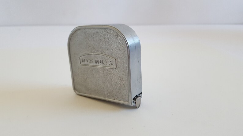 Puede incluir: Un flex&oacute;metro de metal plateado con cuerpo rectangular y parte superior redondeada. El texto "MADE IN U.S.A." est&aacute; grabado en la parte delantera. Herramienta de medici&oacute;n de distancias.
