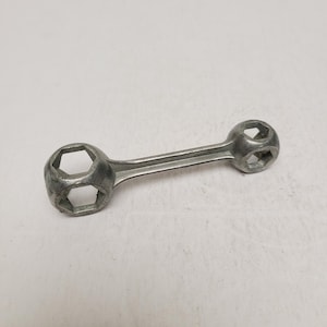 May include: A silver-colored metal multi-socket wrench featuring a central handle bar connecting two circular ends. Each end contains multiple hexagonal openings of varying sizes designed to fit different bolt heads for manual mechanical tasks.