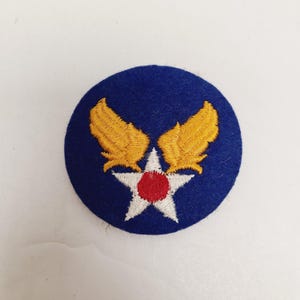 May include: A circular embroidered patch featuring a deep blue background with a central white five-pointed star. The star has a solid red circle in its center and is flanked by two golden yellow wings extending upwards from the star's points.