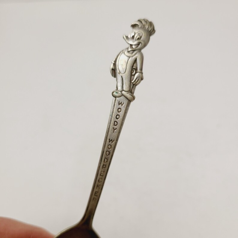 May include: A vintage silver spoon featuring a detailed figure of Woody Woodpecker at the top. The spoon's handle is inscribed with "WOODY WOODPECKER" in capital letters. The spoon has a classic design and is likely a collectible item.