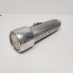 May include: A vintage silver flashlight with the word "HOMART" printed on the side. The flashlight has a cylindrical body with a circular lens and a switch on top. The metal has a worn appearance.
