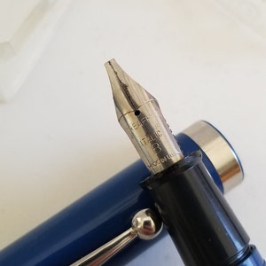 May include: Close-up of a blue fountain pen with a silver nib. The nib is marked "ITALIC" and "MADE IN U.S.A.". The pen has a silver clip and a black section where the nib is attached.
