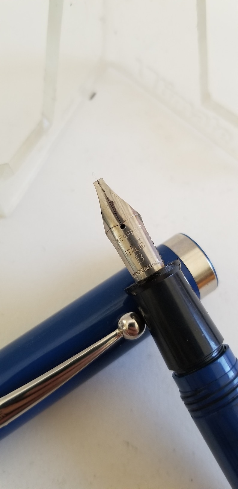May include: Close-up of a blue fountain pen with a silver nib. The nib is marked "ITALIC" and "MADE IN U.S.A.". The pen has a silver clip and a black section where the nib is attached.