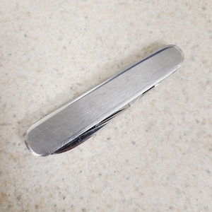 May include: A closed multi-tool pocket knife featuring a brushed silver-tone metal handle with rounded ends. The compact, rectangular tool has a smooth, metallic finish and a visible hinge mechanism along the side, showing a folded blade edge.