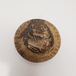 May include: A round, bronze-colored medallion with raised relief figures. The central image depicts a person holding a child. The outer edge features the words "ENTIA MEDICINA ANTIQVE SALVTARI". The medallion has a weathered appearance.