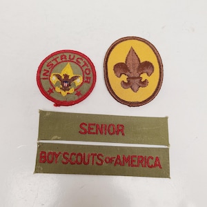 May include: A collection of vintage Boy Scouts of America patches and badges. Includes a round Instructor patch, a fleur-de-lis patch, and two olive green name tapes embroidered with "SENIOR" and "BOY SCOUTS OF AMERICA" in red.