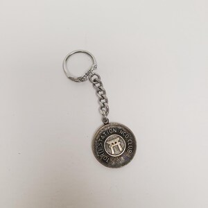 May include: A silver-colored keychain with a circular pendant. The pendant features the text "TORII STATION NCO CLUB" around the edge and a graphic of a torii gate in the center. The keychain has a chain and a ring.
