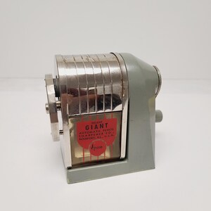 May include: A vintage Apsco Deluxe Giant automatic pencil sharpener with a sage green metal base and a chrome-plated cylindrical housing. The front features a red label with black text reading Deluxe Giant Automatic Pencil Sharpener Co.