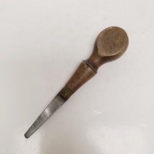 May include: A vintage awl tool with a wooden handle and a metal blade. The handle is oval-shaped and dark brown, with a lighter brown section near the blade. The blade is a flat, rectangular shape and tapers to a point.