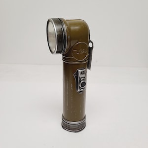 May include: An olive drab right-angle flashlight with a cylindrical body and a silver-toned metal bezel. The side of the casing features the embossed text TL-22-A and a rectangular metal switch plate with a black button for operation.