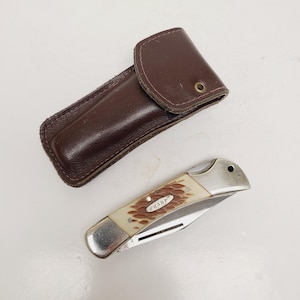 May include: A brown leather knife sheath next to a folding knife. The knife has a silver blade and a cream-colored handle with the word "SHARP" printed on it. The sheath is rectangular with a flap.