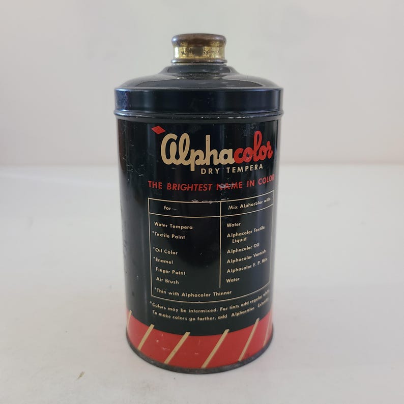 May include: A black tin can with a red and gold label. The label reads "Alphacolor Dry Tempera The Brightest Name in Color". The can contains dry tempera paint.