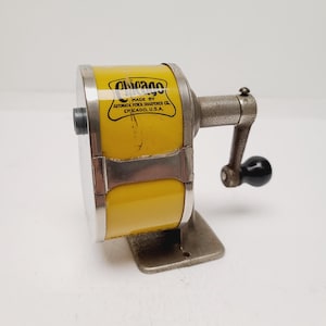 May include: A vintage Chicago pencil sharpener with a bright yellow cylindrical body and a textured gray metal base and crank handle. The sharpener features a black handle knob and a label reading Chicago Made by Automatic Pencil Sharpener Co.