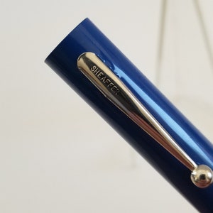May include: Close-up of a blue Sheaffer pen. The pen has a silver clip with the brand name "SHEAFFER" engraved on it. The pen is held at an angle, with the tip facing left.