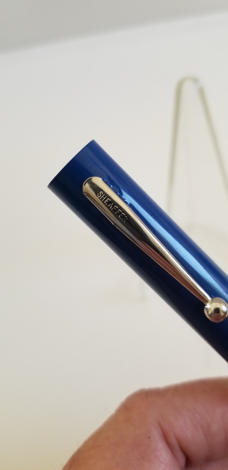 May include: Close-up of a blue Sheaffer pen. The pen has a silver clip with the brand name "SHEAFFER" engraved on it. The pen is held at an angle, with the tip facing left.