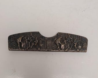 May include: A silver-toned metal object with a curved shape and a central cutout. The surface features a detailed relief depicting figures engaged in various activities, possibly a historical scene. The object has a dark, aged appearance.