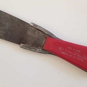 May include: A red handled putty knife with a metal blade. The handle has the text "The Wolverine" and "Union, N.J. U.S.A." printed on it. The handle also has a circular logo with a wolf's head inside.