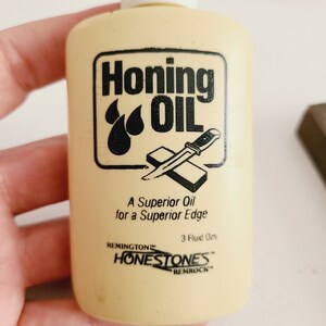 May include: A small, yellow plastic bottle of honing oil with a black label that reads "Honing Oil" and features a knife being sharpened on a honing stone. The label also says "A Superior Oil for a Superior Edge" and "Remington Honestones Remrock".
