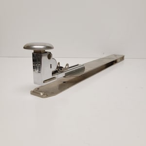 May include: A vintage, chrome-plated stapler with a long base. The stapler features a large, rounded top and a lever mechanism. The base is rectangular and has a slightly aged appearance. The stapler is designed for heavy-duty use.