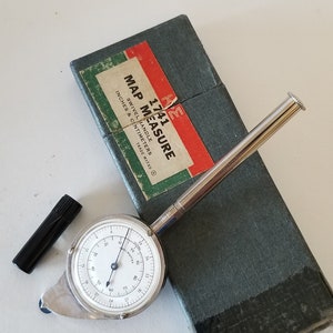 May include: A silver map measure with a swivel handle, measuring in inches and centimeters. The tool is in its original box, which is green with a red label that reads "1741 Map Measure, Swivel Handle, Inches & Centimeters, Trade Marks ®".