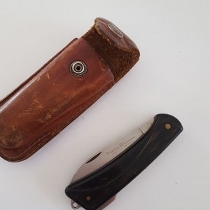 May include: A black and silver folding knife with a black handle and a brown leather sheath. The knife is open, revealing the blade. The sheath is rectangular with a snap closure.