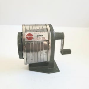 May include: A silver and gray metal pencil sharpener with a crank handle. The sharpener has a red label that reads "Apsco products inc. GIANT Las Angeles, Calif Rockford Ill, USA Type 3A Cutter Assembly".