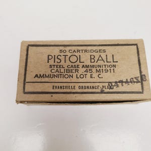 May include: A rectangular cardboard box labeled "50 CARTRIDGES PISTOL BALL STEEL CASE AMMUNITION CALIBER .45 M1911 AMMUNITION LOT E. C." The box is tan with black text and a stamped number. The text "EVANSVILLE ORDNANCE PLANT" is also visible.